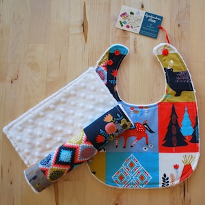 May include: A multi-colored baby bib with a white trim and a white, soft, textured burp cloth. The bib has a patchwork design with a bear, trees, and geometric patterns. The bib has two red buttons on the sides. The burp cloth has a white, soft, textured surface.