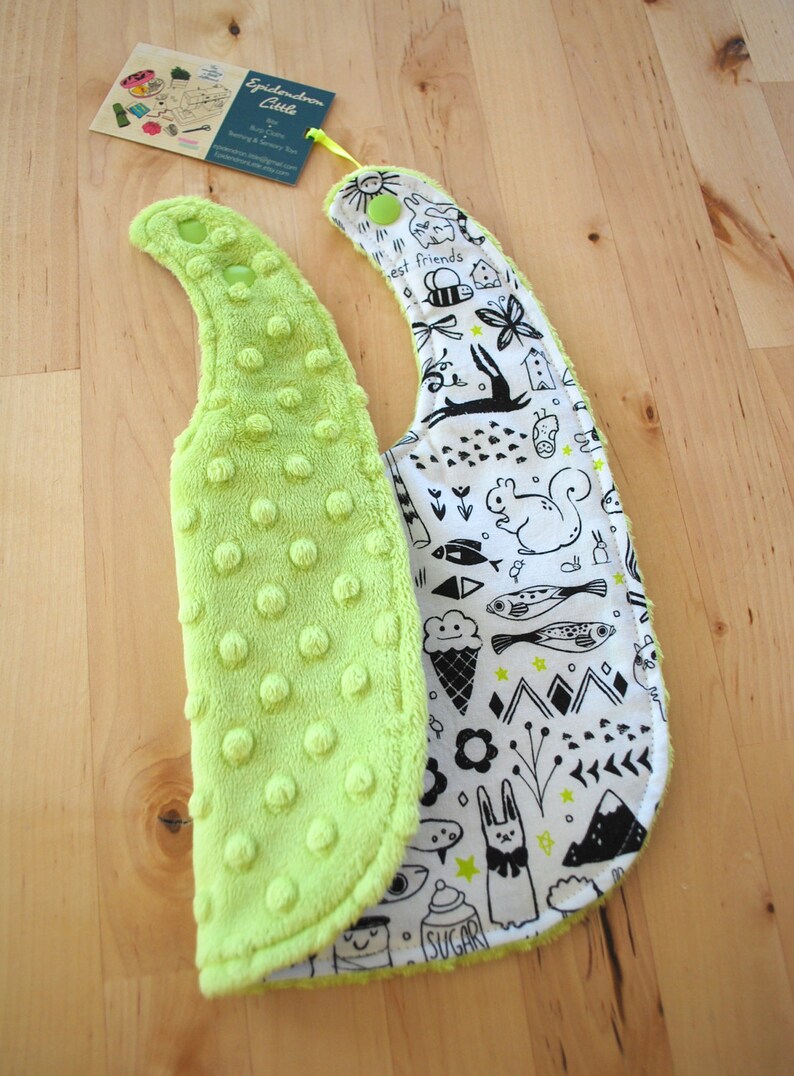 May include: A reversible baby bib with a lime green soft, textured side and a white side with black and white illustrations of animals, stars, and other designs. The text "best friends" is visible on the white side.