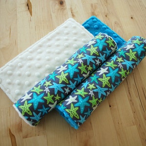 May include: Two rolled up fabric crib bumpers with a blue and green starfish pattern on a gray background. The bumpers are on top of a white fabric with small white dots.