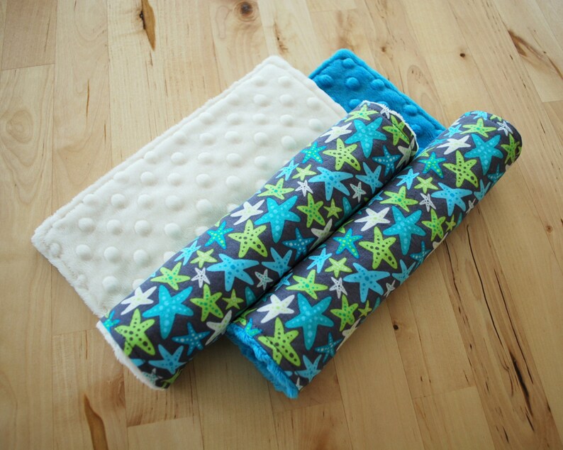 May include: Two rolled up fabric crib bumpers with a blue and green starfish pattern on a gray background. The bumpers are on top of a white fabric with small white dots.