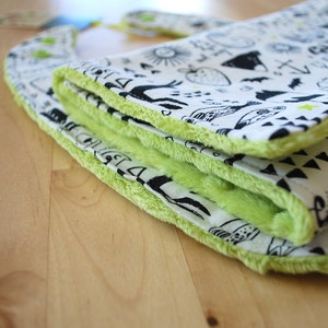 May include: A green and white baby bib with a black and white print of various designs. The bib is folded and has a soft, fuzzy texture.