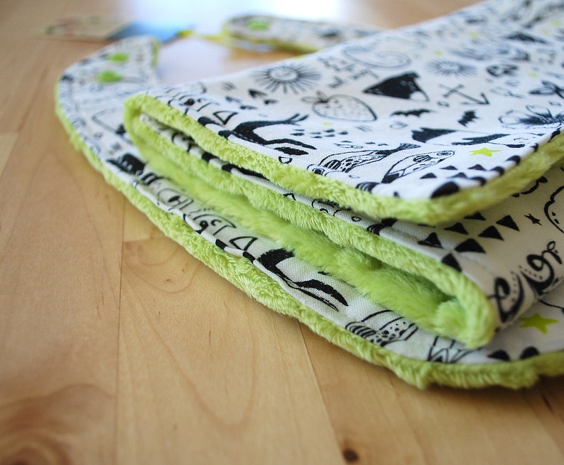 May include: A green and white baby bib with a black and white print of various designs. The bib is folded and has a soft, fuzzy texture.
