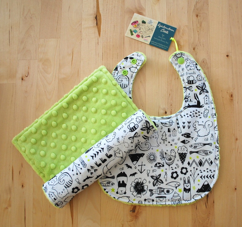 May include: A green and white baby bib with a black and white print of animals and nature elements. The bib is made of fabric and has a soft, textured feel. The bib is paired with a green and white baby burp cloth with a similar print.
