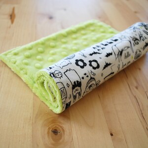 May include: A green and white baby blanket with a playful black and white pattern. The blanket is rolled up and features a variety of illustrations including animals, flowers, and geometric shapes. The text "hello" and "i love you" is visible on the blanket.