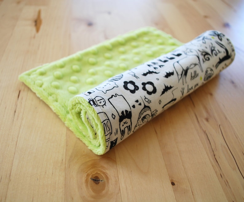 May include: A green and white baby blanket with a playful black and white pattern. The blanket is rolled up and features a variety of illustrations including animals, flowers, and geometric shapes. The text "hello" and "i love you" is visible on the blanket.