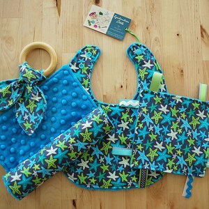 May include: A blue and green baby bib, a blue and green baby blanket, a blue and green baby toy, and a wooden teething ring. The items are all decorated with a starfish pattern.