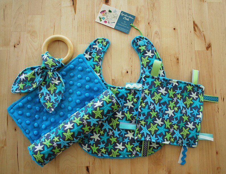 May include: A blue and green baby bib, a blue and green baby blanket, a blue and green baby toy, and a wooden teething ring. The items are all decorated with a starfish pattern.