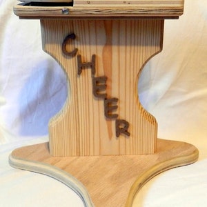 Könnte beinhalten: Holzener Cheerleading-Ständer mit dreieckigem Sockel und zwei Plattformen. Der Ständer ist aus hellem Holz gefertigt und hat das Wort "CHEER" auf der Vorderseite.