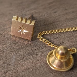 Puede incluir: Pasador de corbata dorado con diseño de estrella y cadena. El pasador tiene forma rectangular con un diseño de estrella en el centro. La cadena está unida a un respaldo dorado.