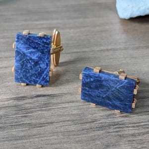 May include: A pair of cufflinks featuring rectangular, deep blue stones with gold-colored metal accents. The stones have a marbled appearance, and the cufflinks are displayed on a wooden surface.