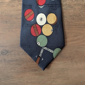 May include: Navy blue necktie featuring a pattern of billiard balls in various colors, including red, green, and yellow. The tie also has a cue stick design at the bottom. The tie is a classic accessory for a formal or casual outfit.