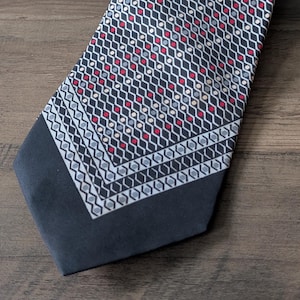 May include: A necktie featuring a dark charcoal background with a repeating geometric pattern of white zig-zag lines and small red and light gray dots. The bottom section of the tie has a solid dark charcoal border with a white geometric trim.