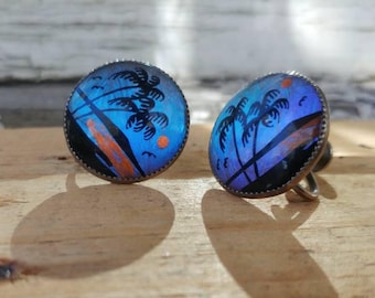 May include: A pair of blue and orange butterfly wing earrings with a silver setting. The earrings feature a tropical scene with palm trees and a sunset.