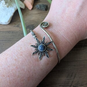 May include: A silver-toned metal cuff bracelet featuring a spiral end and a sun-shaped charm. The sun charm has a dark blue circular stone at its center with radiating points and small dot details. The bracelet has an open, adjustable design.