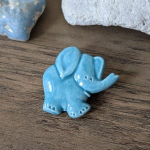 Puede incluir: Pequeña figura de elefante de cerámica azul claro con acabado brillante. El elefante tiene detalles, incluyendo trompa, orejas y pies. La figura está colocada sobre una superficie de madera, con otras piedras decorativas en el fondo.