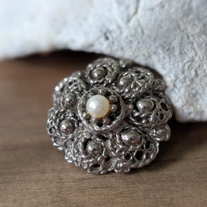 May include: A silver-toned, vintage brooch with a floral design. The brooch features a central, round, off-white pearl surrounded by intricate, textured metalwork. The brooch is set against a wooden surface and a white rock.