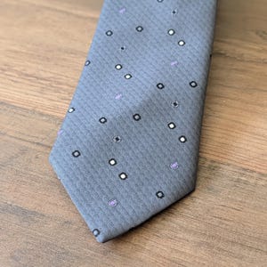May include: A grey necktie featuring a subtle diagonal herringbone weave pattern. The fabric is decorated with a repeating motif of small black squares, white dots, and light purple circular accents scattered across the surface of the tie.