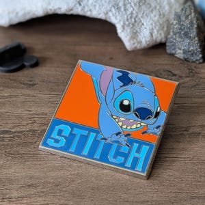 以下が含まれることがあります： リロ＆スティッチのキャラクター、スティッチが描かれた正方形のエナメルピン。オレンジと青の背景に青いスティッチのデザインがあり、「STITCH」の文字が青いブロック体で書かれています。