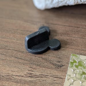 以下が含まれることがあります： 長方形のトップと2つの円形の耳が付いた、黒いミッキーマウス型のピンバック。ピンは木製の表面にあり、背景には白いオブジェクトがあり、ミッキーマウスをテーマにした銀色のデザインがあります。