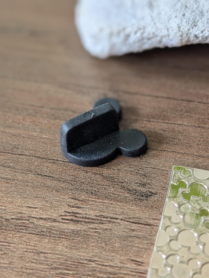 以下が含まれることがあります： 長方形のトップと2つの円形の耳が付いた、黒いミッキーマウス型のピンバック。ピンは木製の表面にあり、背景には白いオブジェクトがあり、ミッキーマウスをテーマにした銀色のデザインがあります。