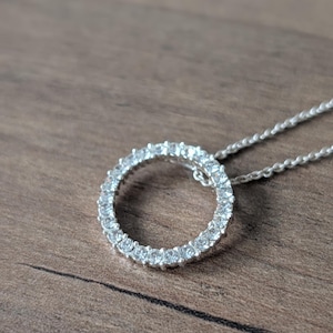 Peut inclure: Un collier argenté avec un pendentif circulaire orné de pierres claires et scintillantes. Le pendentif est suspendu à une fine chaîne. Le bijou est posé sur une surface en bois.