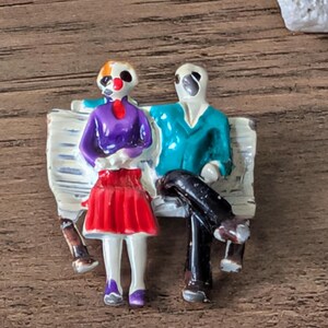 May include: A painted figurine of a couple sitting on a bench. The woman wears a purple top and red skirt, while the man has a teal jacket. The bench is white with a distressed look. The figurine is on a wooden surface.