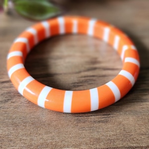 Puede incluir: Brazalete de rayas naranja y blancas. El brazalete tiene forma circular, con bandas alternas de color naranja brillante y blanco. Hecho de un material brillante, descansa sobre una superficie de madera.