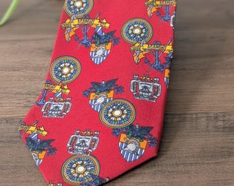 May include: A red tie with a repeating pattern of navy blue anchors, compasses, and crests. The crests have a white and blue shield design. The tie is made of a smooth fabric and is displayed on a wooden surface.