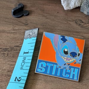 以下が含まれることがあります： リロ＆スティッチのキャラクター、スティッチが描かれた正方形のエナメルピン。青とオレンジの配色です。ピンのサイズは約2.5cmで、「STITCH」の文字が青いブロック体で書かれています。