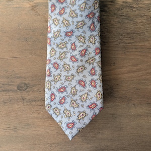 May include: A pale blue tie featuring a repeating paisley pattern in red, yellow, and blue. The tie is made of a smooth fabric and is displayed on a wooden surface. The paisley design is detailed and evenly spaced.