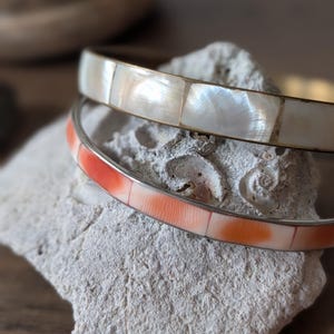 May include: Two bangle bracelets. One is white with rectangular mother-of-pearl inlays, and the other is orange and white with rectangular inlays. Both have silver-coloured metal bands. The bracelets are displayed on a textured, light-coloured stone.