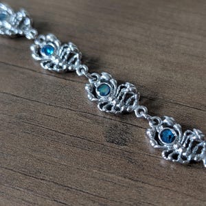 May include: A silver-toned bracelet with an ornate design. Each link features a detailed, openwork pattern surrounding a small, round, blue gemstone. The bracelet is displayed on a wooden surface, highlighting its intricate craftsmanship.