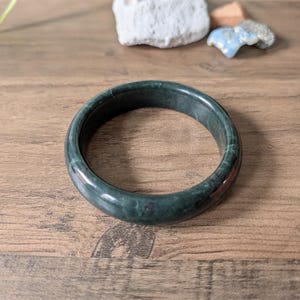 May include: A dark green jade bangle bracelet. The smooth, polished surface of the bracelet reflects light, showcasing its natural variations. The circular bracelet is a classic jewelry piece.