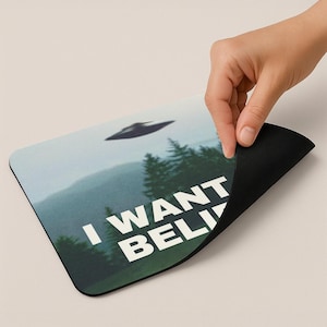 May include: A black and white mousepad with a green, blue, and white image of a flying saucer over a forest. The text "I WANT TO BELIEVE" is printed on the mousepad.