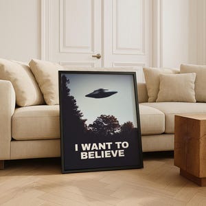 May include: A black framed poster with a photo of a flying saucer over a forest. The poster has the text "I WANT TO BELIEVE" in white letters.