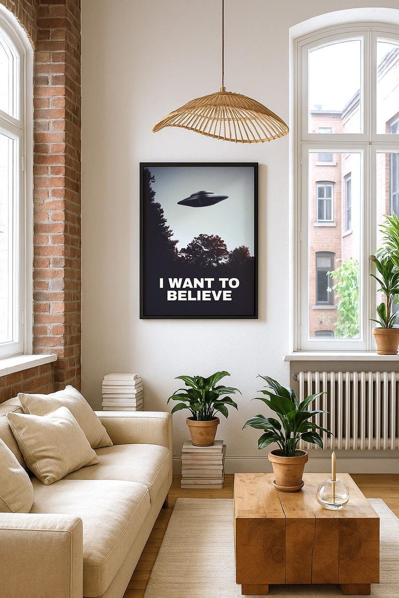 May include: A living room interior with a beige sofa, a wooden coffee table, and two potted plants. A framed poster with the text "I WANT TO BELIEVE" and a flying saucer is hanging on the wall. A woven pendant light hangs from the ceiling.