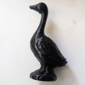 May include: A small, black, three-dimensional printed figurine of a goose standing on a circular base. The surface of the goose displays visible horizontal layer lines characteristic of the additive manufacturing process, with a smooth, matte finish.