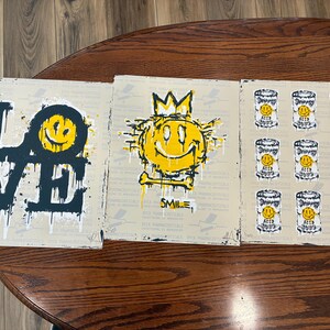 May include: Three black and white art prints with yellow accents. The first print says "LOVE" in bold black letters. The second print features a yellow smiley face with a crown and the word "SMILE". The third print has nine yellow smiley faces in cans.
