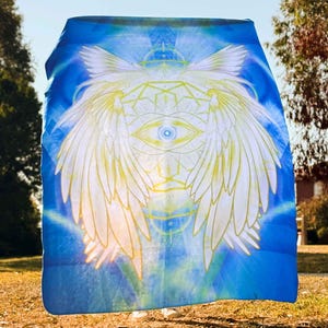 May include: A blue blanket with a celestial design featuring a central eye, wings, and geometric patterns. The design is in white and yellow against a blue background. The blanket is held outdoors in natural light.