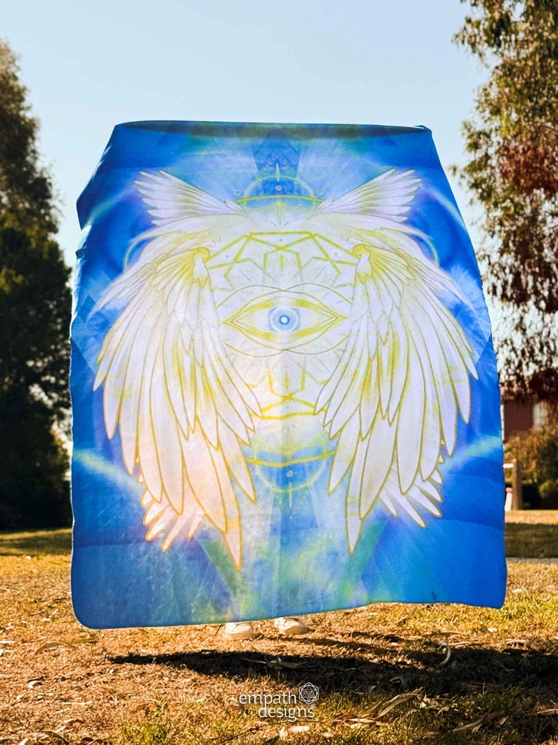 May include: A blue blanket with a celestial design featuring a central eye, wings, and geometric patterns. The design is in white and yellow against a blue background. The blanket is held outdoors in natural light.