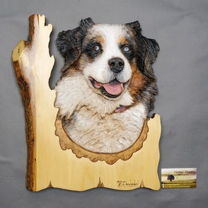 Può includere: Una scultura in legno di una testa di cane, con pelo marrone, bianco e nero. La scultura è inserita in un pezzo di legno che sembra un tronco d'albero. La firma dell'artista è visibile nell'angolo in basso a destra.