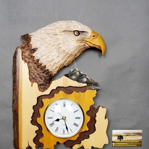 May include: A wooden wall clock carved in the shape of an eagle's head with a clock face in the center. The eagle is carved in a realistic style with brown and white feathers. The clock face is white with black Roman numeral markings. The clock is mounted on a wooden base with a brown and light wood finish. The clock is signed "DavydovArt" and includes the website address "www.etsy.com/ca/shop/DavydovArt".