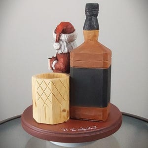Puede incluir: Figurita de madera de Pap&aacute; Noel sosteniendo un vaso de ponche de huevo, sentado junto a una botella de whisky de madera. La figurita y la botella est&aacute;n sobre una base de madera con la inscripci&oacute;n "V. Darlyden".