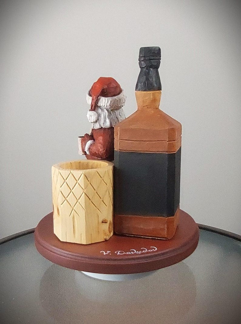 Puede incluir: Figurita de madera de Pap&aacute; Noel sosteniendo un vaso de ponche de huevo, sentado junto a una botella de whisky de madera. La figurita y la botella est&aacute;n sobre una base de madera con la inscripci&oacute;n "V. Darlyden".