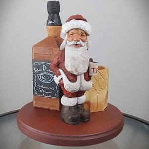 Puede incluir: Una figurita de madera de Pap&aacute; Noel sosteniendo una taza peque&ntilde;a, de pie junto a una botella de whisky y un barril de madera. La botella tiene una etiqueta que dice "Jack Daniel's No. 1 Whiskey".