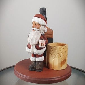 Puede incluir: Una figura de madera de Pap&aacute; Noel sosteniendo una taza que dice "TEA" y de pie junto a una botella de madera y un portavasos de madera. La figura est&aacute; sobre una base redonda de madera.