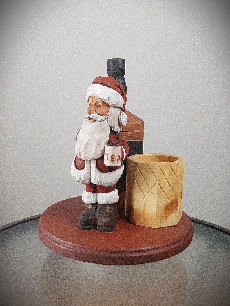 Puede incluir: Una figura de madera de Pap&aacute; Noel sosteniendo una taza que dice "TEA" y de pie junto a una botella de madera y un portavasos de madera. La figura est&aacute; sobre una base redonda de madera.