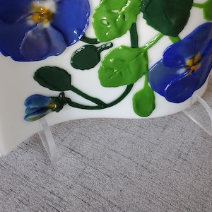 Op de afbeelding: Een wit glazen paneel met een blauw en groen bloemenontwerp. Het paneel is gemonteerd op een transparante acrylstandaard.