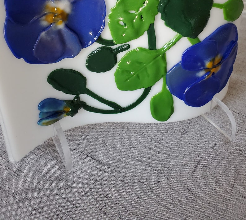 Op de afbeelding: Een wit glazen paneel met een blauw en groen bloemenontwerp. Het paneel is gemonteerd op een transparante acrylstandaard.