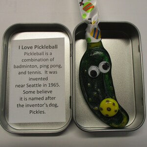 Peut inclure: Une petite d&eacute;coration en verre en forme de cornichon vert avec des yeux globuleux et un charme de balle de pickleball jaune. La d&eacute;coration est dans une bo&icirc;te en &eacute;tain argent&eacute;e avec une carte blanche qui dit "J'aime le pickleball. Le pickleball est une combinaison de badminton, de ping-pong et de tennis. Il a &eacute;t&eacute; invent&eacute; pr&egrave;s de Seattle en 1965. Certains pensent qu'il porte le nom du chien de l'inventeur, Pickles."